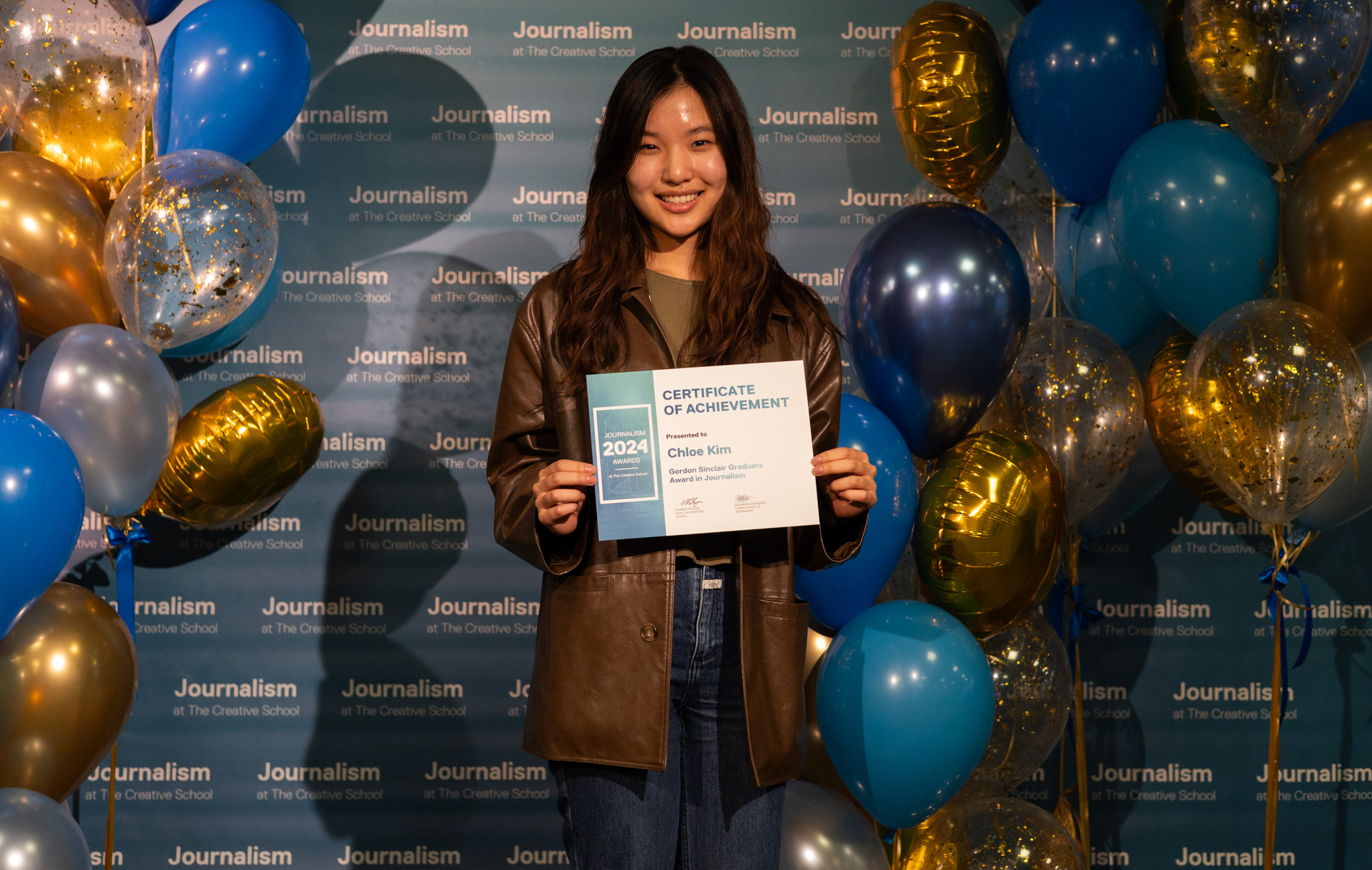 Chloe Kim stands in front of Journalism at The Creative School backdrop with balloons on each side while holding a certificate.